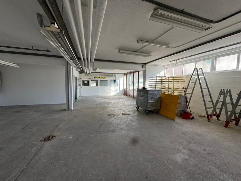 Empty warehouse with white walls, concrete floor, a few windows, multiple ceiling lights, a few scattered items like a trash can, buckets, a yellow board, and ladders
