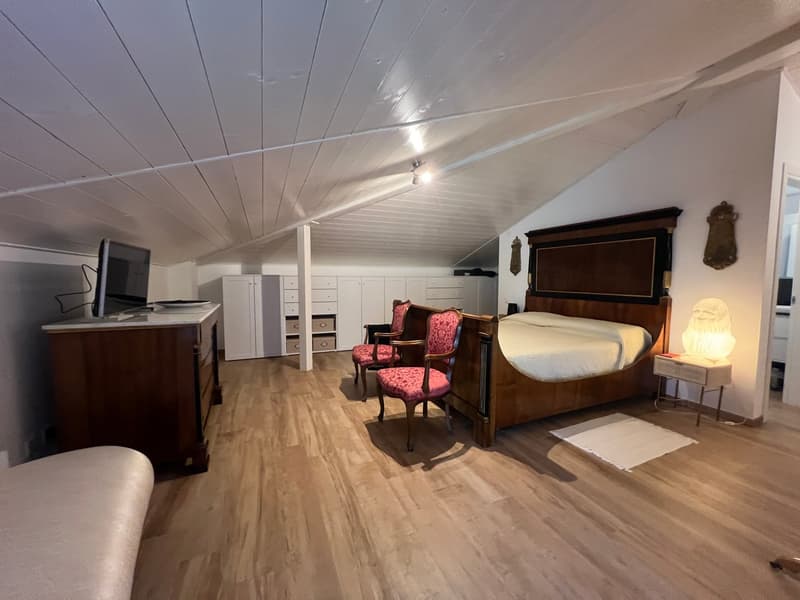 Bedroom with wooden floor, white walls, large bed, two chairs, desk with monitor, lamp, and cabinets