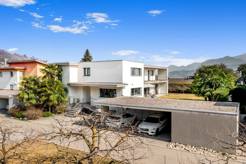 Two-story modern house with a flat roof, surrounded by palm trees and other vegetation. The house has a garage and parking spaces underneath. The exterior is a mix of white and gray colors.