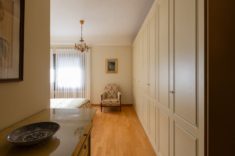 wooden floor, white walls, large wardrobe, bed, chair, chandelier, window with curtains, decorative bowl on table