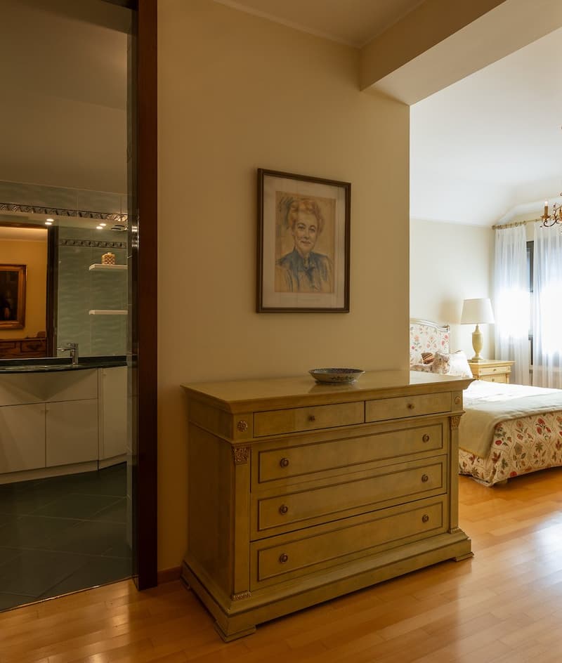 bedroom with wooden floor, bed, nightstand, lamp, picture frame, wooden dresser, bathroom with sink and mirror