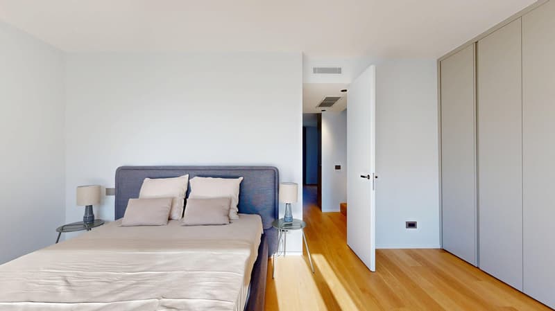 Spacious bedroom with a large bed, gray upholstered headboard, and wooden flooring. The room has a clean, minimalist design with white walls and a doorway leading to another room.