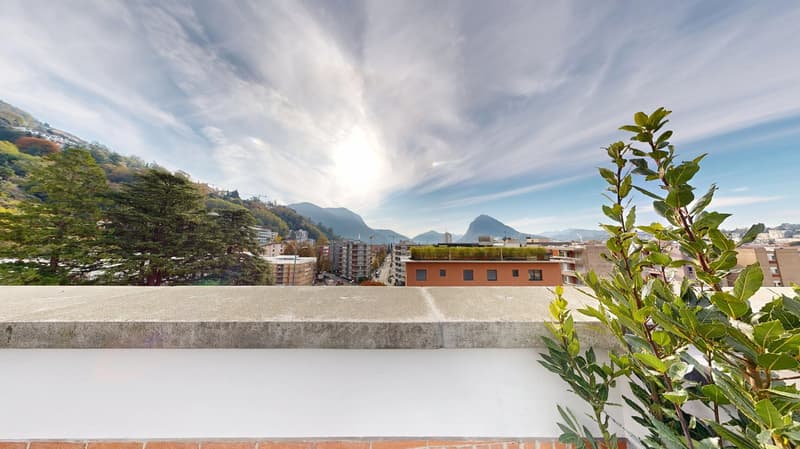 The image shows a panoramic view of a city surrounded by mountains. There are several multi-story residential and commercial buildings visible, with a balcony or terrace on one of the buildings in the foreground. The sky is partly cloudy, creating an atmo