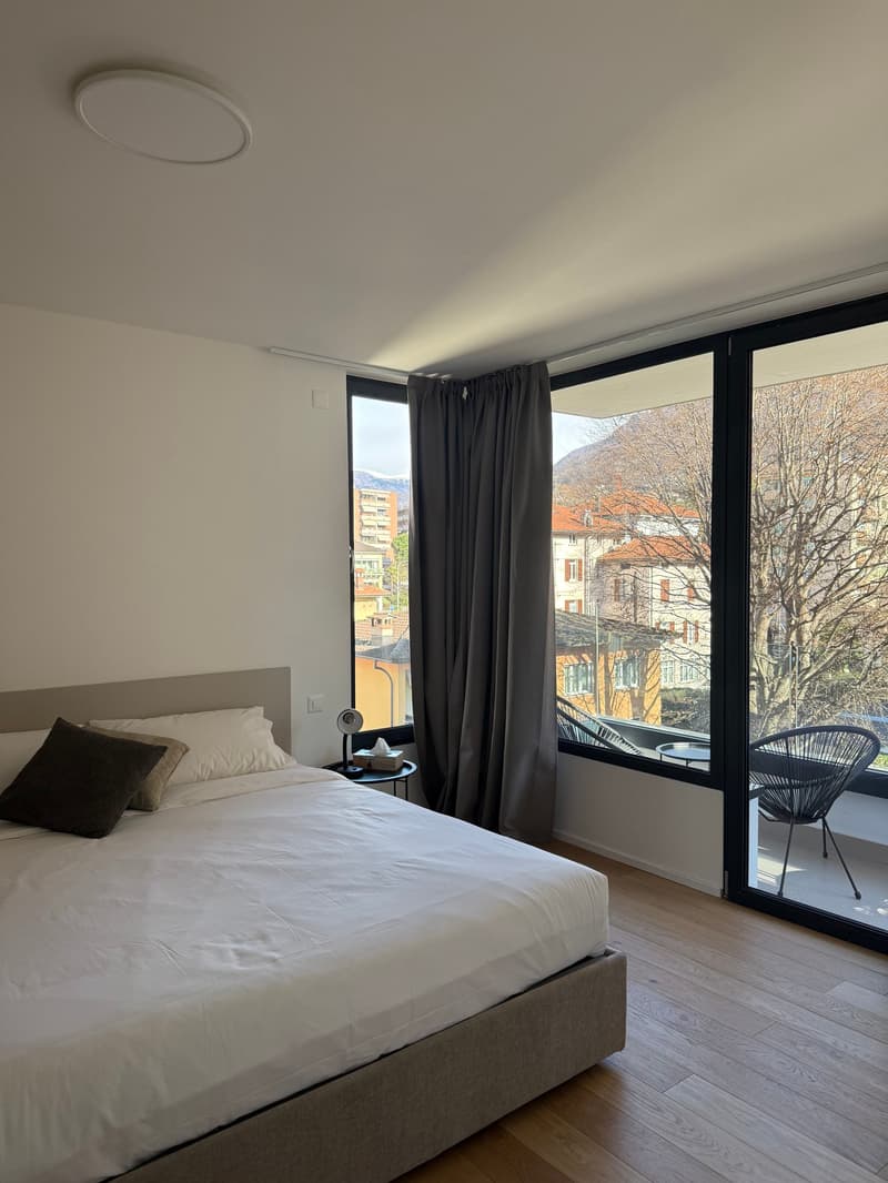a bed with a white duvet, nightstand, pillows, large windows with curtains