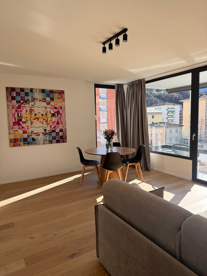 Dining area with wooden table, chairs, and artwork. Open to living area with couch and glass doors leading to balcony.
