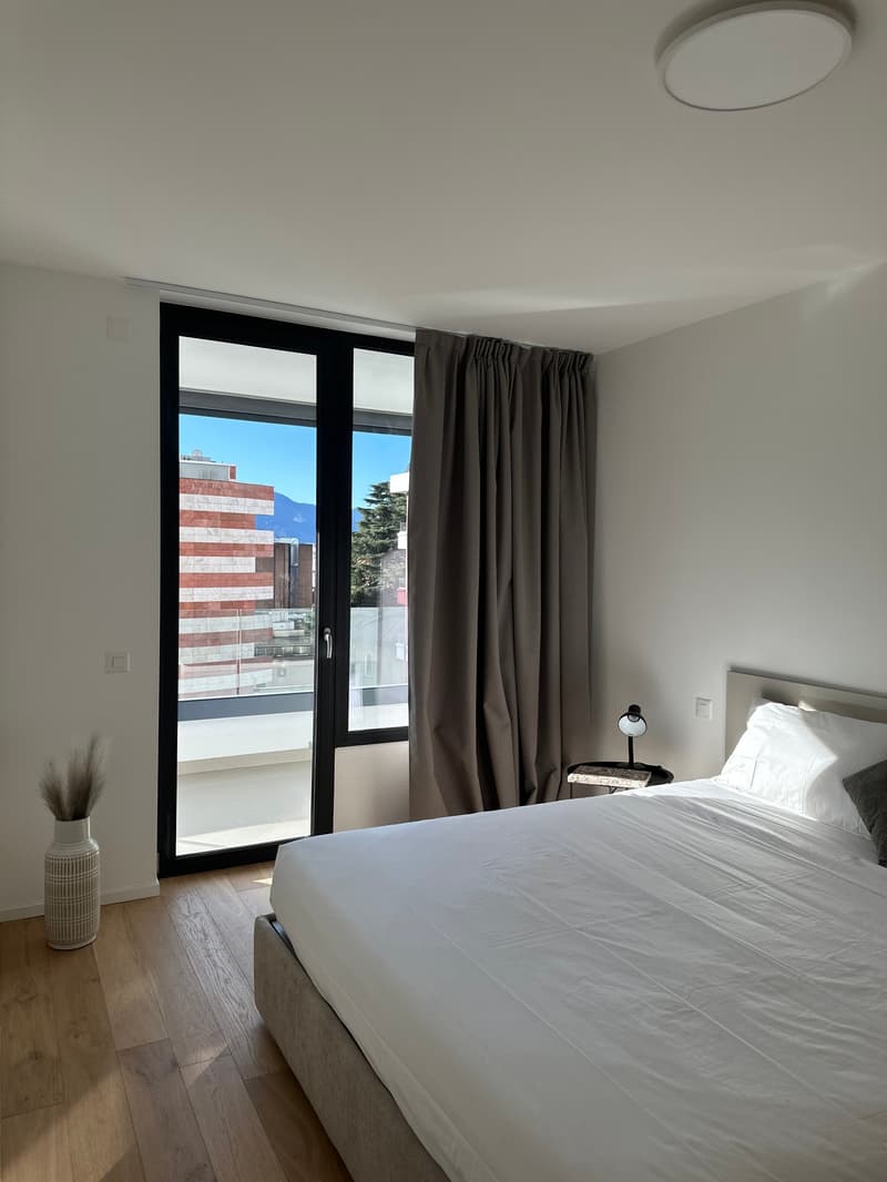 Sliding glass door to the balcony, double bed, lamp on nightstand, vase, hardwood floors, curtains