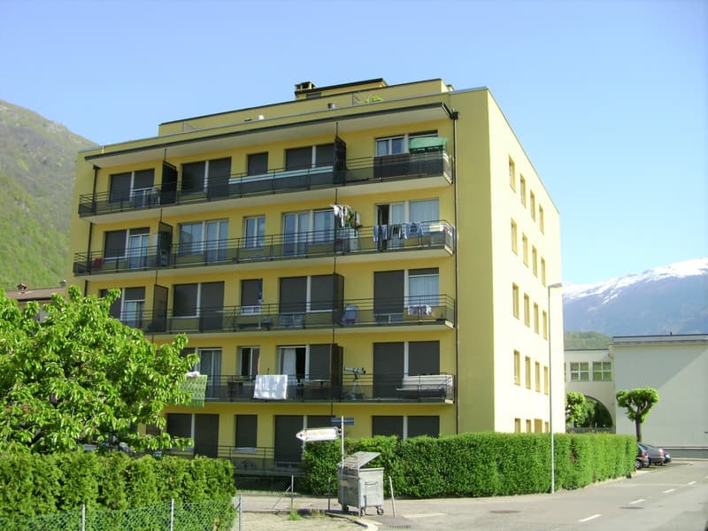 6 story building, yellow exterior, multiple balconies, green awnings on 4th floor balcony, street sign near front entrance, green hedges in the front.