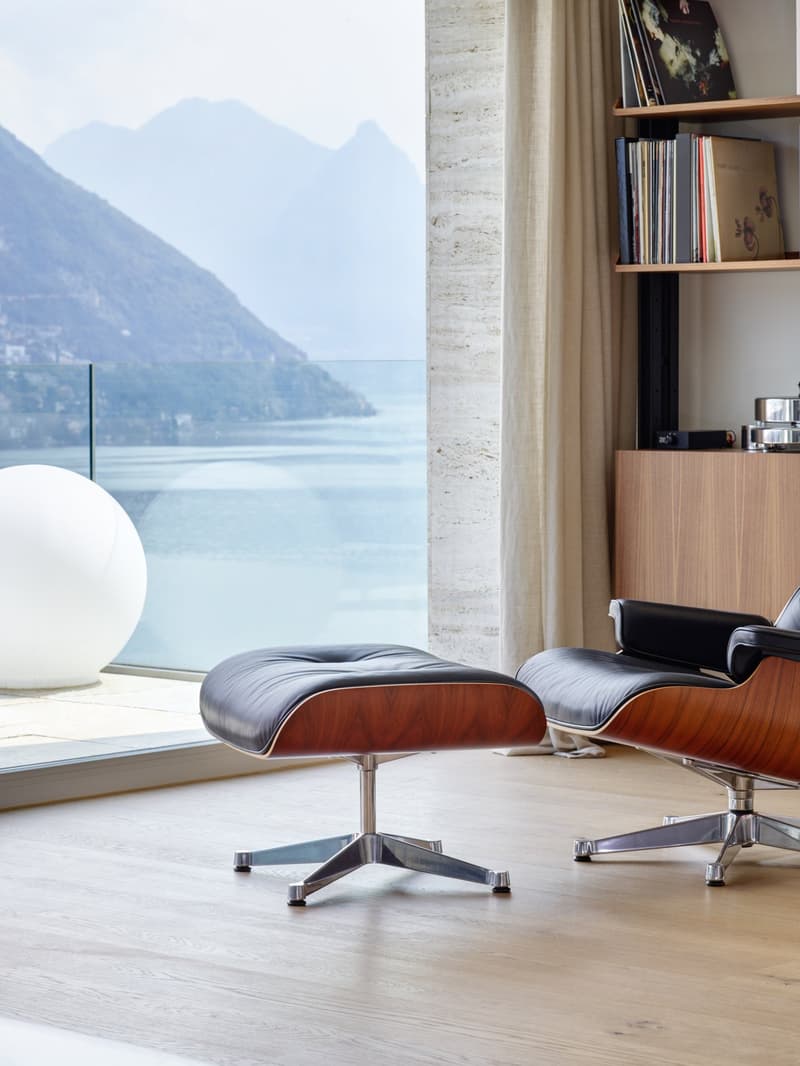 modern living room with large glass window, leather armchair, ottoman, wooden floor, white sphere, bookshelf, records, curtain