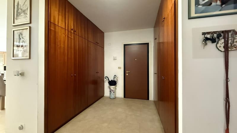 Empty hallway with wooden doors on each side, a closed door at the end, tiled floor, and a picture frame and umbrella on either side of the closed door.