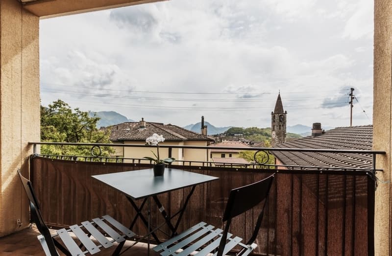 Balcony with a small table and chairs, overlooking a town with a church steeple and mountains in the background