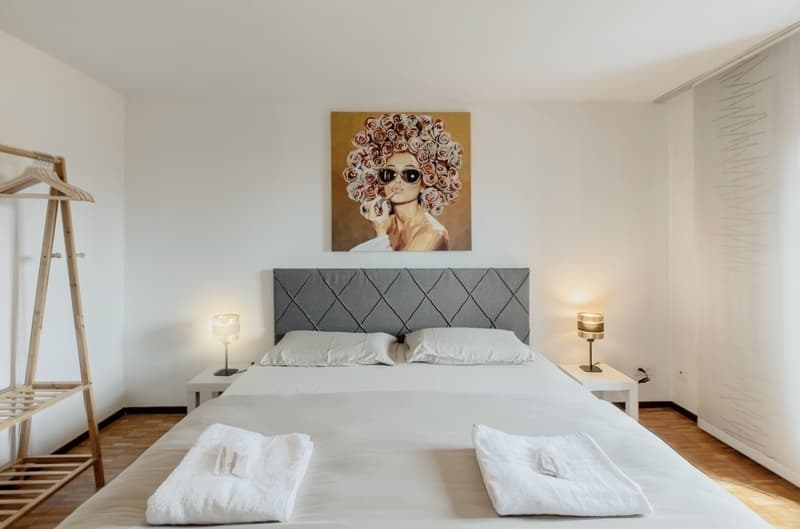 Spacious bedroom with a large bed, gray upholstered headboard, and a painting of a woman with a floral hairstyle on the wall. The room also features wooden floors, bedside tables with lamps, and a clothing rack.