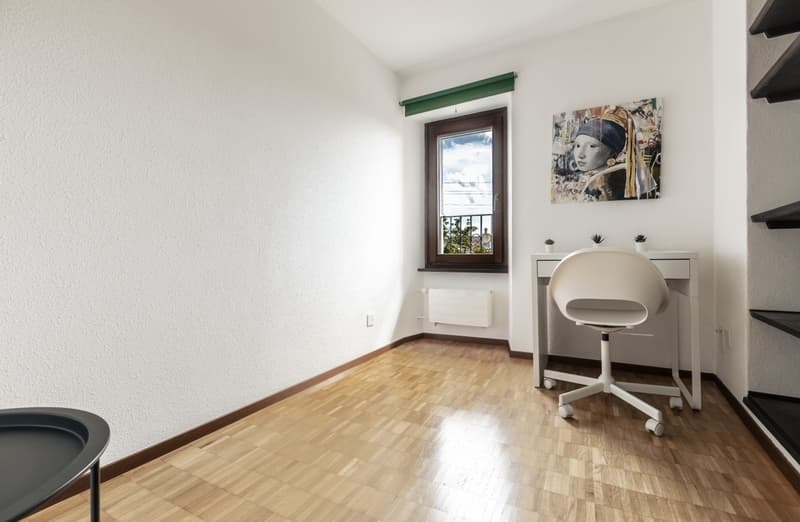 Spacious bedroom with hardwood flooring, white walls, a large window, and a desk with a chair. The room also features shelving and a decorative artwork on the wall.