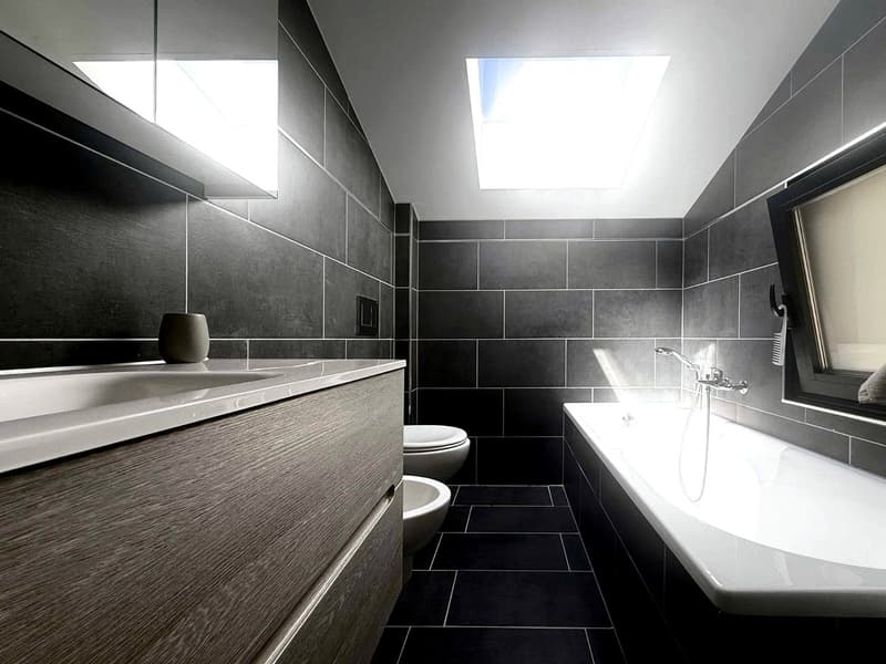 modern bathroom with gray tiles, white bathtub, white sink, two toilets, skylights