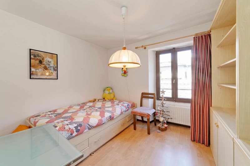 Single bed, wooden chair, wooden shelves, hanging lamp, framed picture, small Christmas tree, window with white curtain, radiator