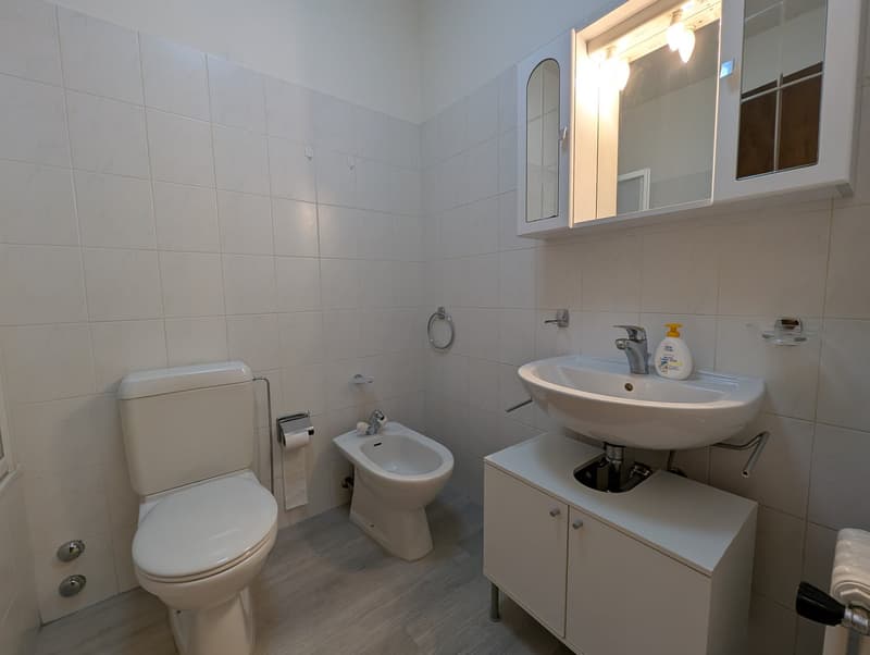 Modern bathroom with white tiles, a bidet, a toilet, a sink, and a cabinet with a mirror.