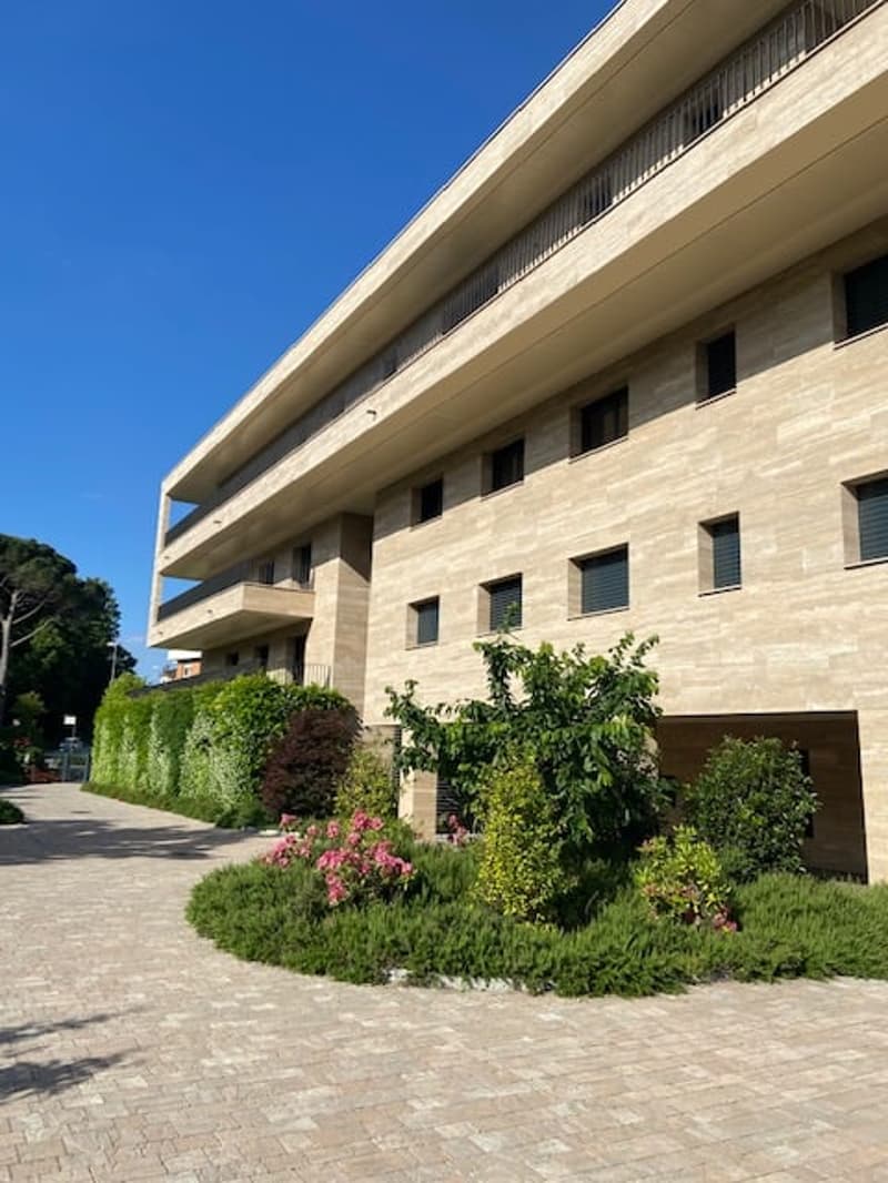 Modern building with multiple floors, beige stone exterior, multiple windows, balconies, driveway, greenery