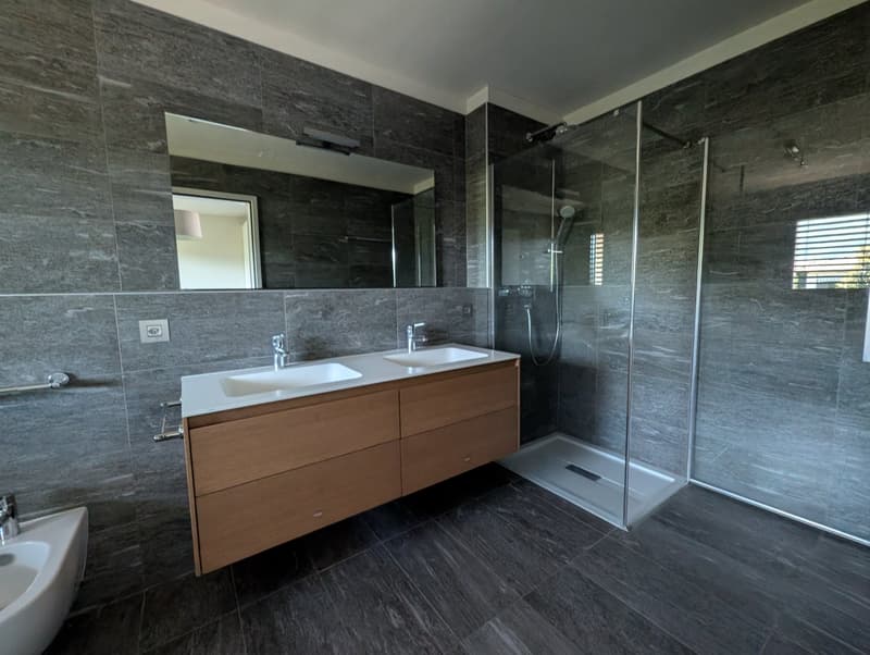 modern bathroom with double sink, large mirror, glass shower enclosure, wooden cabinet, gray tiles