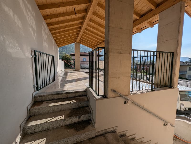 Stone steps leading to a balcony with wooden beams and metal railings, overlooking a street with a parked car and a building.
