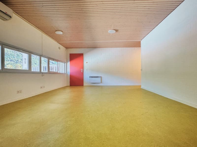 Large open space with wooden ceiling, white walls, and a red door. The room has a concrete floor and several windows providing natural light.