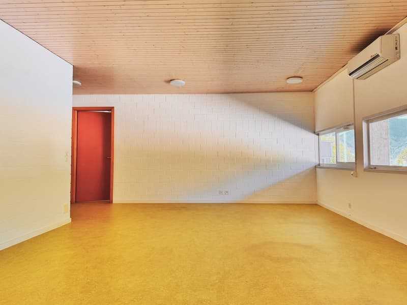 Large open room with white brick walls, wooden ceiling, and yellow concrete flooring. There is a red door and a wall-mounted air conditioning unit visible.