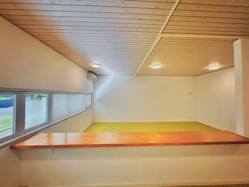 Large open space with wooden ceiling, white brick walls, and a wooden counter or bench along one wall. The room has large windows providing natural light.