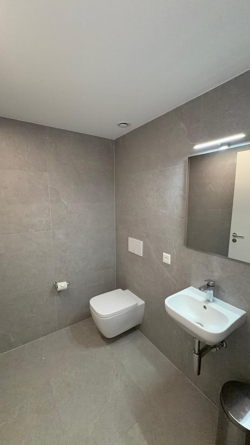 A small bathroom with grey tiled walls, white tiled floor, white toilet, and white sink with silver faucet.