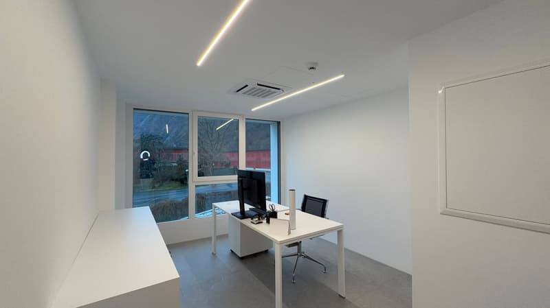 An empty office with a desk, monitor, and office chair in front of a large window, white walls and ceiling, and recessed lighting.