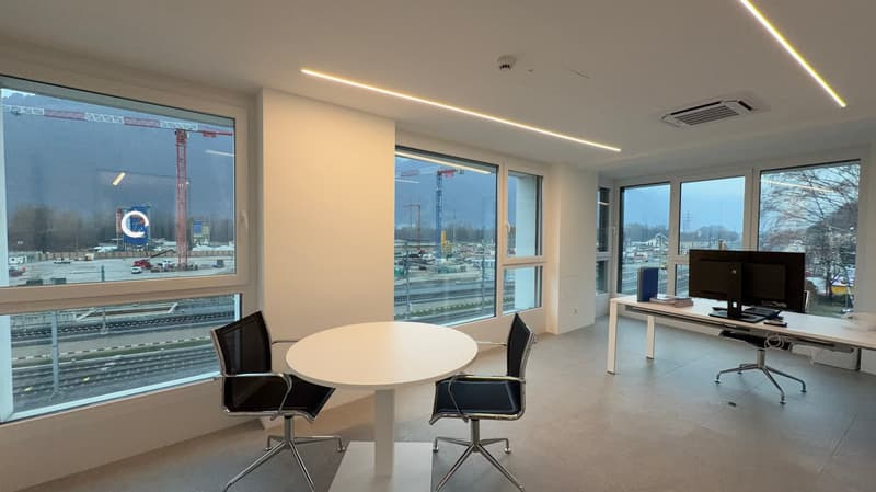 Two black office chairs around a white round table, white desk with computer, office furniture, large windows overlooking train tracks
