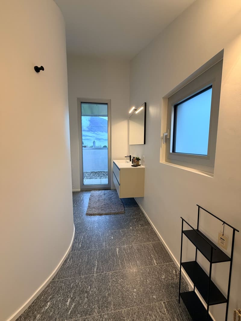Hallway with a curved wall, white tile flooring, large glass window with view, glass door leading to bathroom, modern sink, mirror, black shelf
