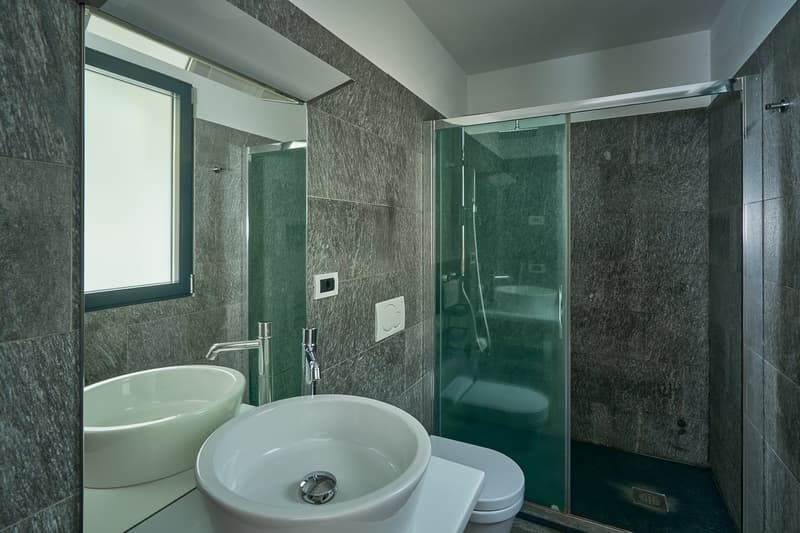 Gray tile walls, white sink, large oval sink, toilet, walk-in shower with glass door, silver fixtures, mirror