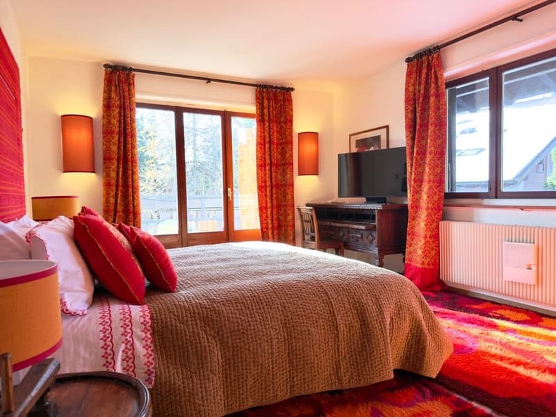 large bed with red pillows, wooden desk with TV, wooden chair, radiator, wooden floor, glass doors, curtains, lamps
