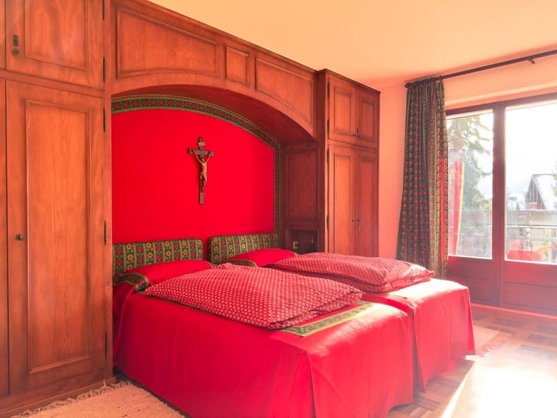 Two twin beds with red sheets and pillows, wooden wardrobes, red wall with a cross, wooden floor, window with curtains