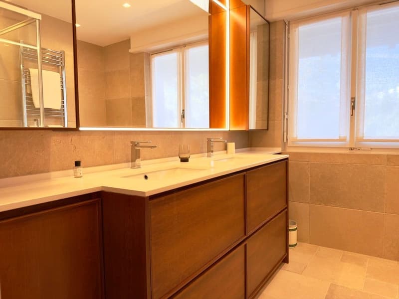 Bathroom with a large mirror, wooden cabinets, double sink, and a shower area.