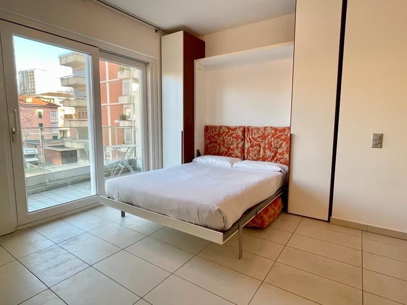 large double bed, white and red bedding, white tiles, sliding glass door to balcony, view of the city, white wardrobe, storage shelf