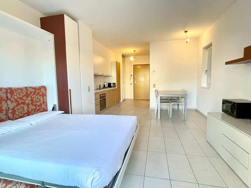 A modern bedroom with a bed, a dining table, and an open-plan kitchen and living area. The kitchen has a microwave, a refrigerator, and a sink. The living area has a couch, a dining table, and a TV. The walls are white, and the floor is tiled. The bedroom