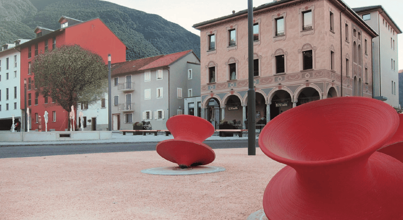 The image shows a town square with colorful buildings, including a pink building with arched windows and a red building. There are also some red sculptural pieces in the foreground of the square.
