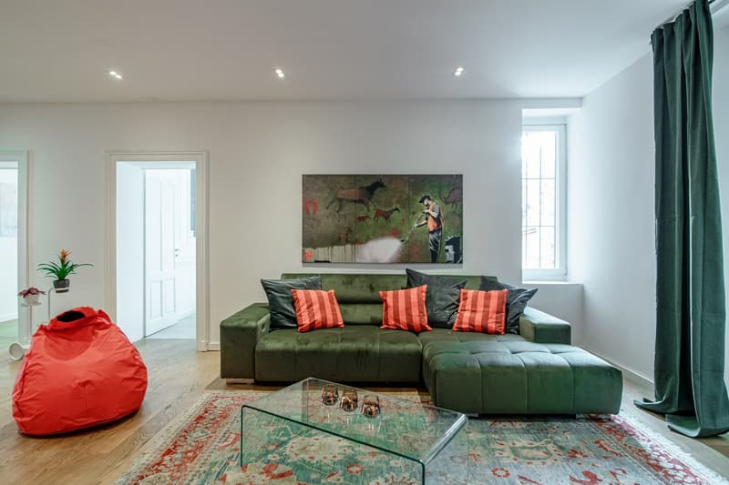 green couch, pillows, glass table, carpet, two small windows, painting on the wall