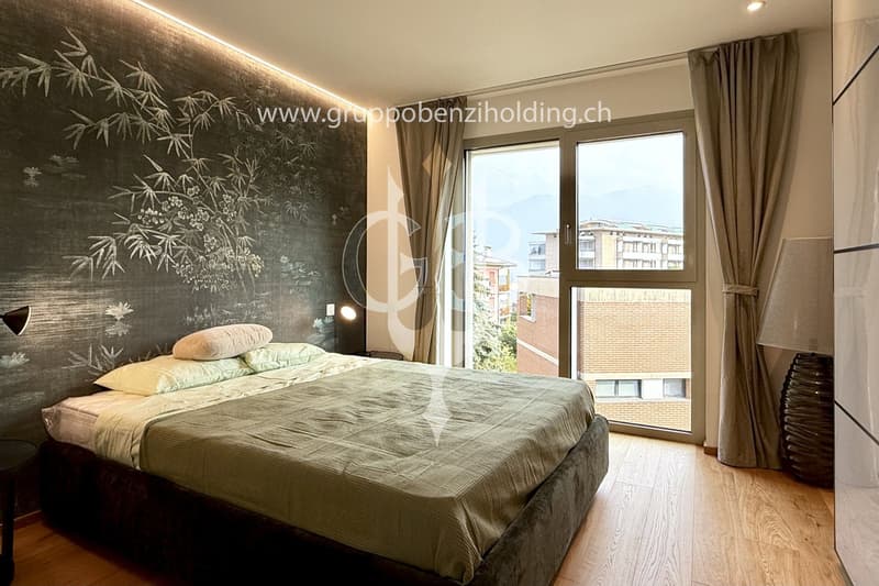 modern double bed, white pillow, beige blanket, dark wooden floor, big glass window, wall lamp, dark gray wall, brown curtains