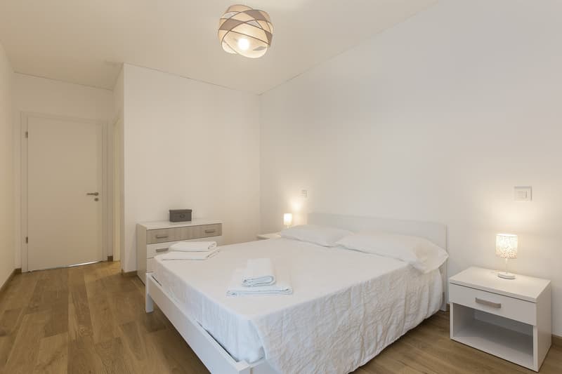 Contemporary bedroom with white walls, wooden floor, double bed with white linen, white nightstands, lamps, and a cabinet with drawers. The room is well-lit with a ceiling light and additional wall lamps.