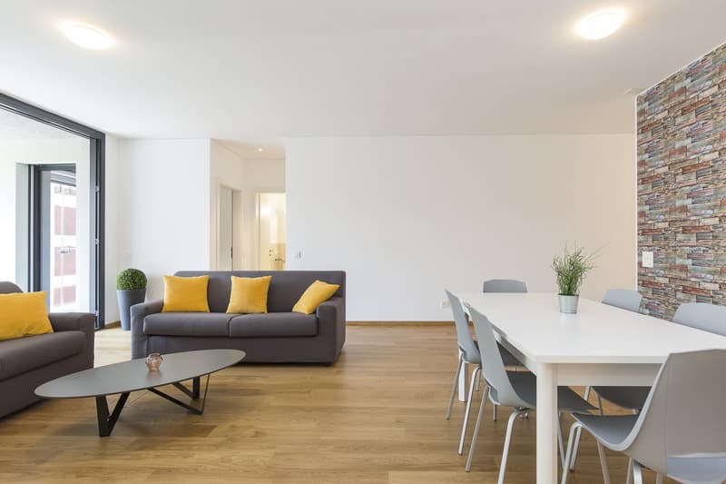 Gray couch, yellow pillows, oval gray coffee table, white dining table, gray chairs, potted plants, white walls, wooden floor, glass wall with doors