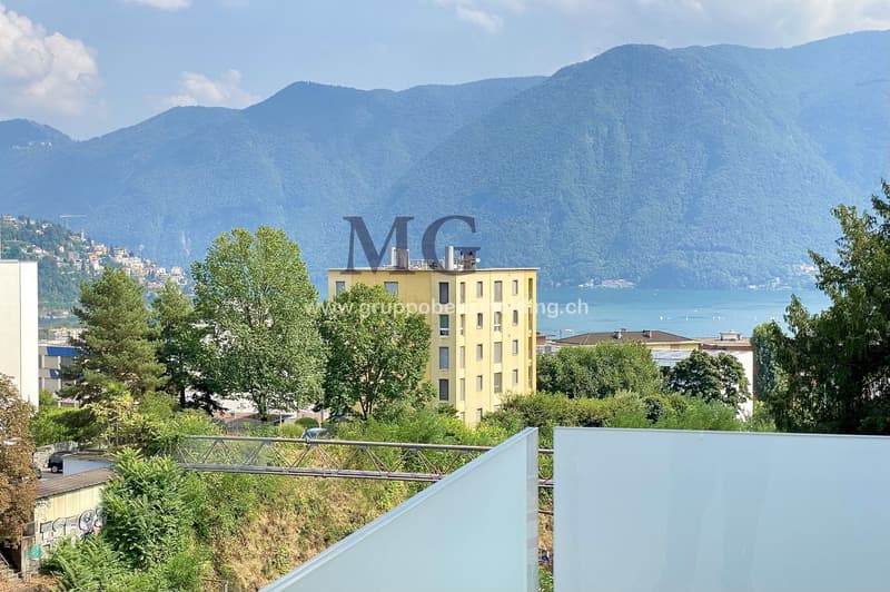 A yellow building with multiple floors, surrounded by greenery and trees. It has a clear view of the mountains and a lake in the distance.