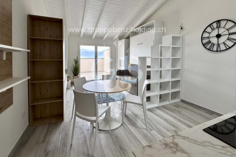 A room with white and gray furniture, including a desk and chairs, a round table, shelves, and a clock on the wall. There is a glass door leading to a balcony with a view of the outdoors.