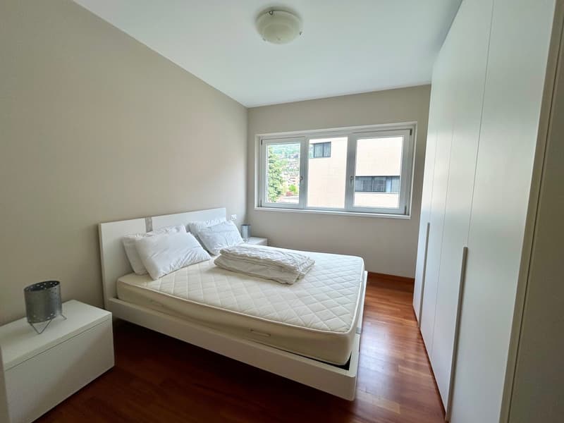 Bedroom with a bed, white walls, wooden floor, large window, white nightstands, white wardrobes, ceiling lamp