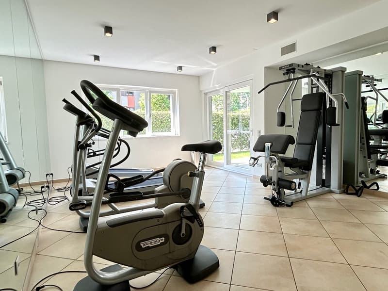 A gym room with various exercise equipment, including treadmills, stationary bikes, and weight machines. The room has white walls, a white ceiling, and beige tiles on the floor. There are windows with glass doors that lead to a grassy area outside.