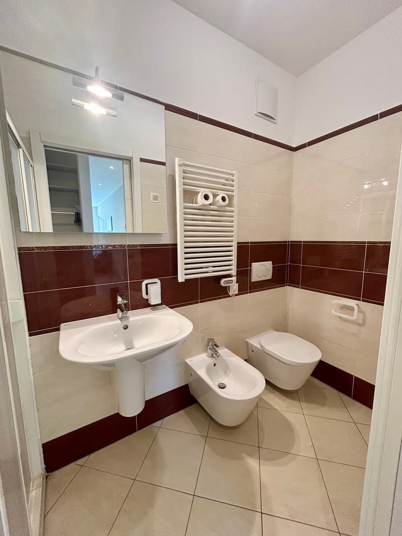 Bathroom with white sink, white bidet, white toilet, mirror, towel rack, tiled floor, brown and white tiles on the wall