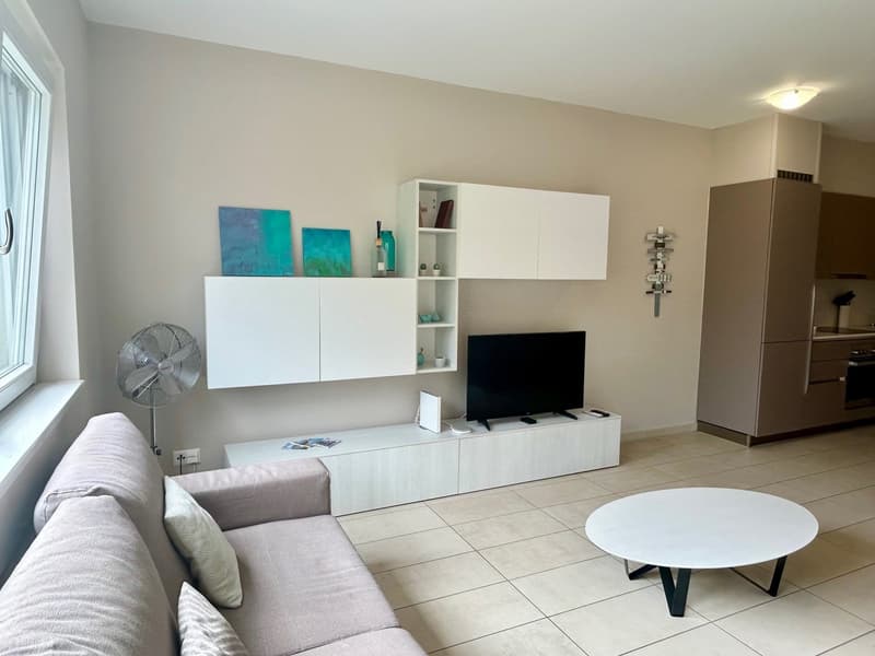Living room with white walls, beige couch, white coffee table, white shelves, flat screen TV, fan, and beige tiled floor.