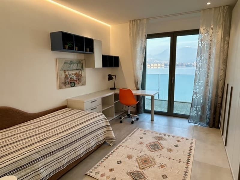 A bedroom with a bed, desk, chair, rug, framed picture, lamps, and windows with a view of the lake and city