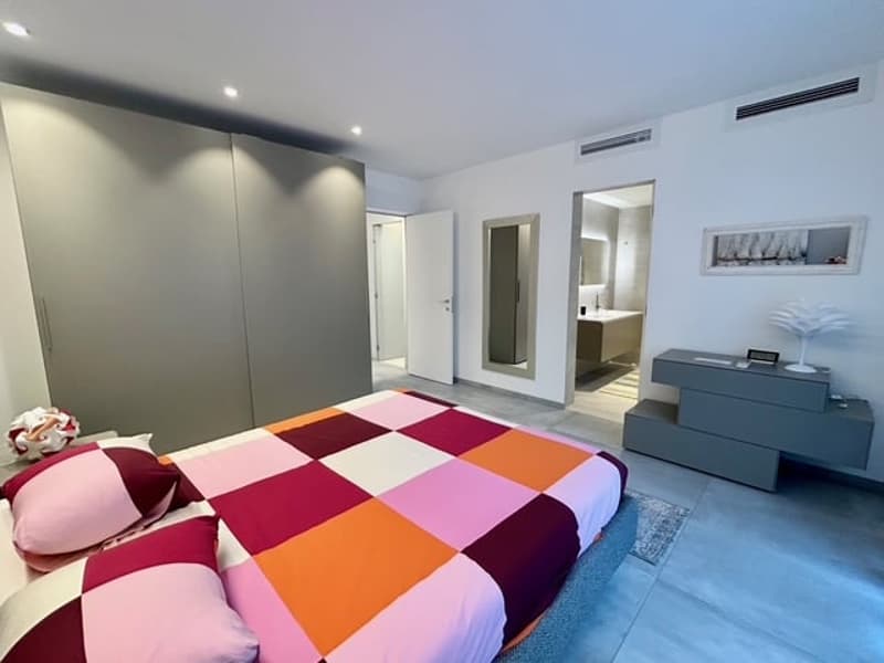 double bed with colorful quilt, pink and maroon pillows, white walls, gray wardrobe, dresser with desk, mirror and picture frame