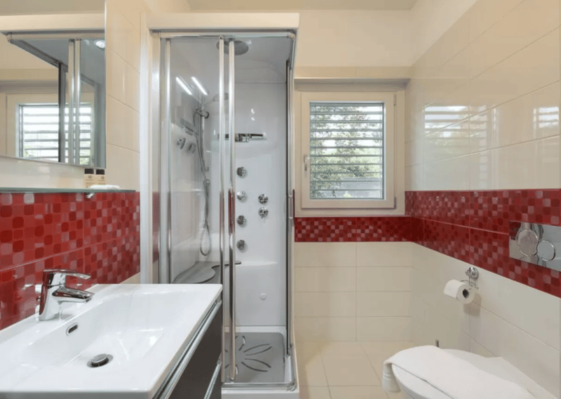 white sink, silver faucet, large glass enclosed shower, white tiles, mirror, toilet, window with jalousie