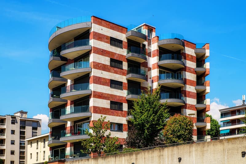 Multi-story apartment building with red brick and glass facade, balconies on each floor, surrounded by trees and other buildings